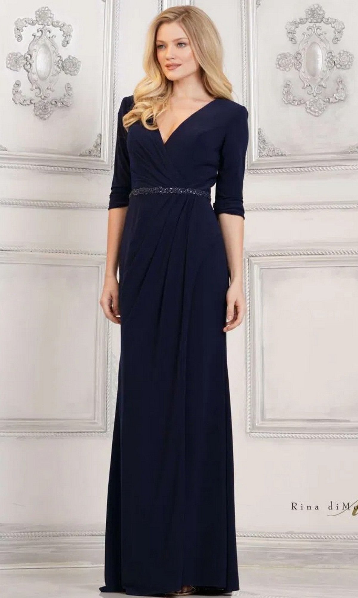 A woman stands before an ornate white wall wearing the Rina Di Montella RD3109, a navy long formal dress with a V-neckline, three-quarter sleeves, matte jersey fabric, and a beaded waist.